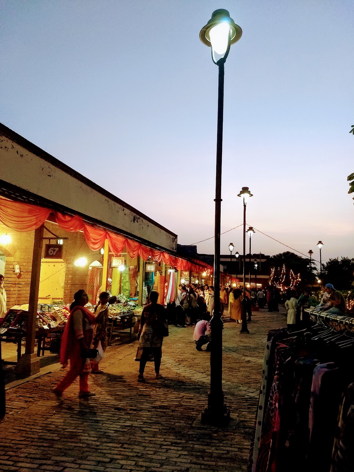 Craft-stall market lanes at Dilli Haat Pitampura at dusk, with numbered stalls 67 to 69 under red drapes and shoppers browsing