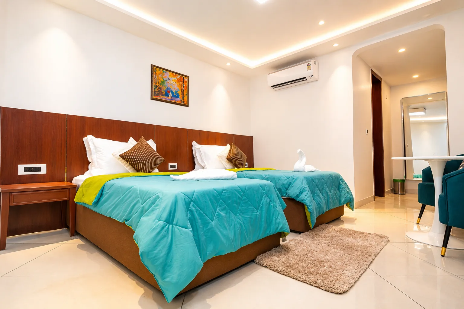 Two twin beds with turquoise throws and swan towels at J60 Suites Delhi, beside a white table and teal armchairs under warm cove lighting.