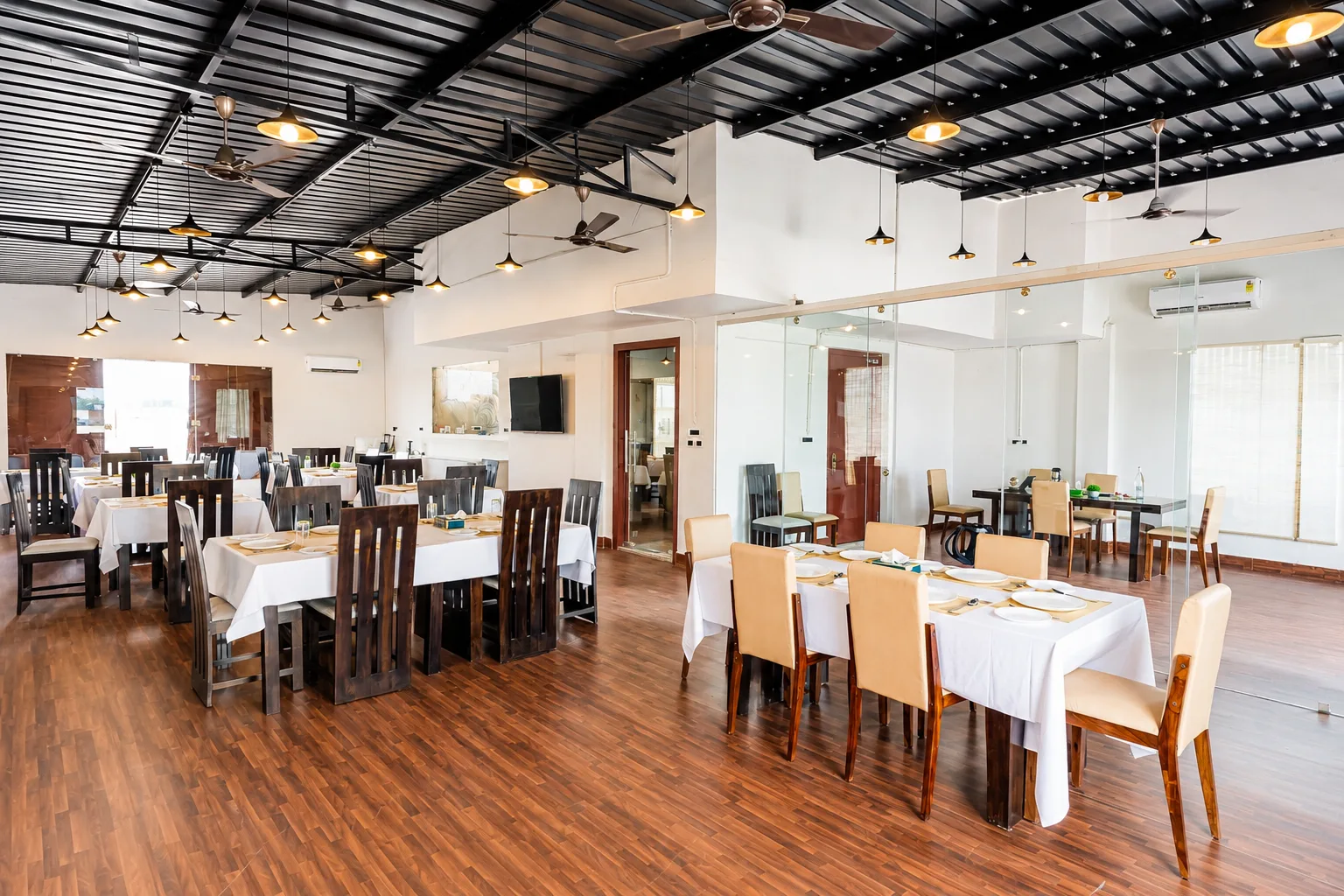 Dining hall at J60 Suites Delhi — white-linen tables under brass pendant lights, warm daylight, and a glass-partitioned private table at right.